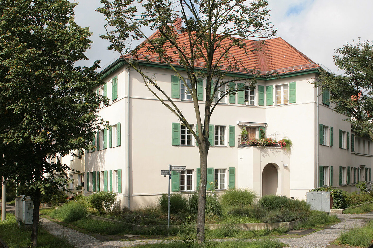An einer Straßenecke steht ein saniertes Wohnhaus mit heller Fassade, grün gestrichenen Fensterläden und rotem Ziegeldach. Darum Pflanzen, Bäume und ein Bürgersteig.