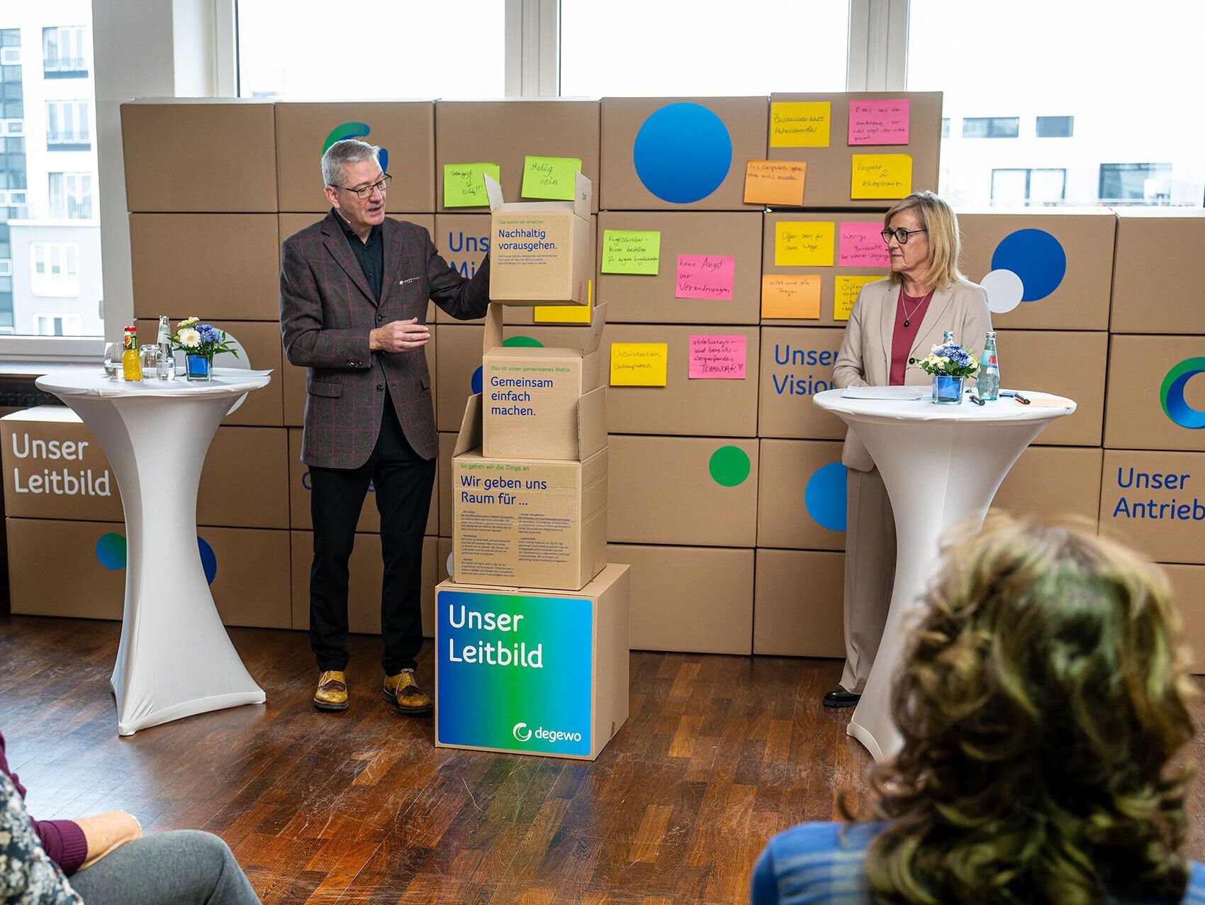 Zwei Personen präsentieren das degewo Leitbild vor einer Wand aus beschrifteten Kartons während sie mit dem Publikum sprechen.