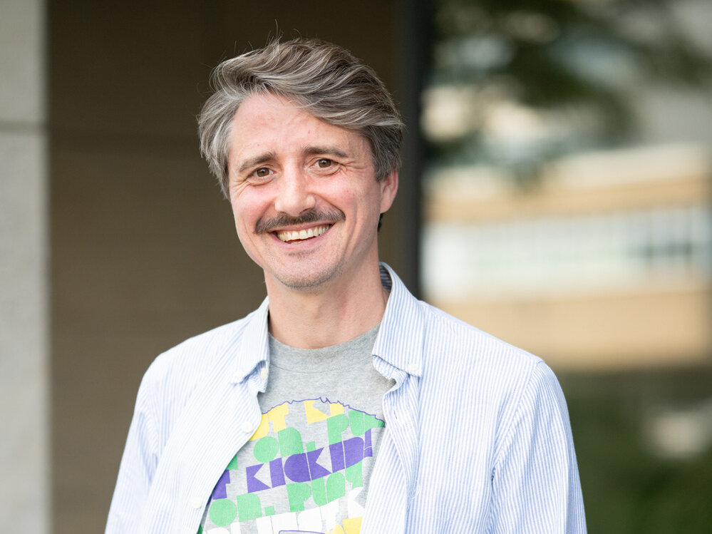 Mann mit grauem Haar und Schnurrbart trägt ein helles Hemd und steht vor einem Gebäude im Freien; er lächelt und blickt in die Kamera, der Hintergrund ist weichgezeichnet.