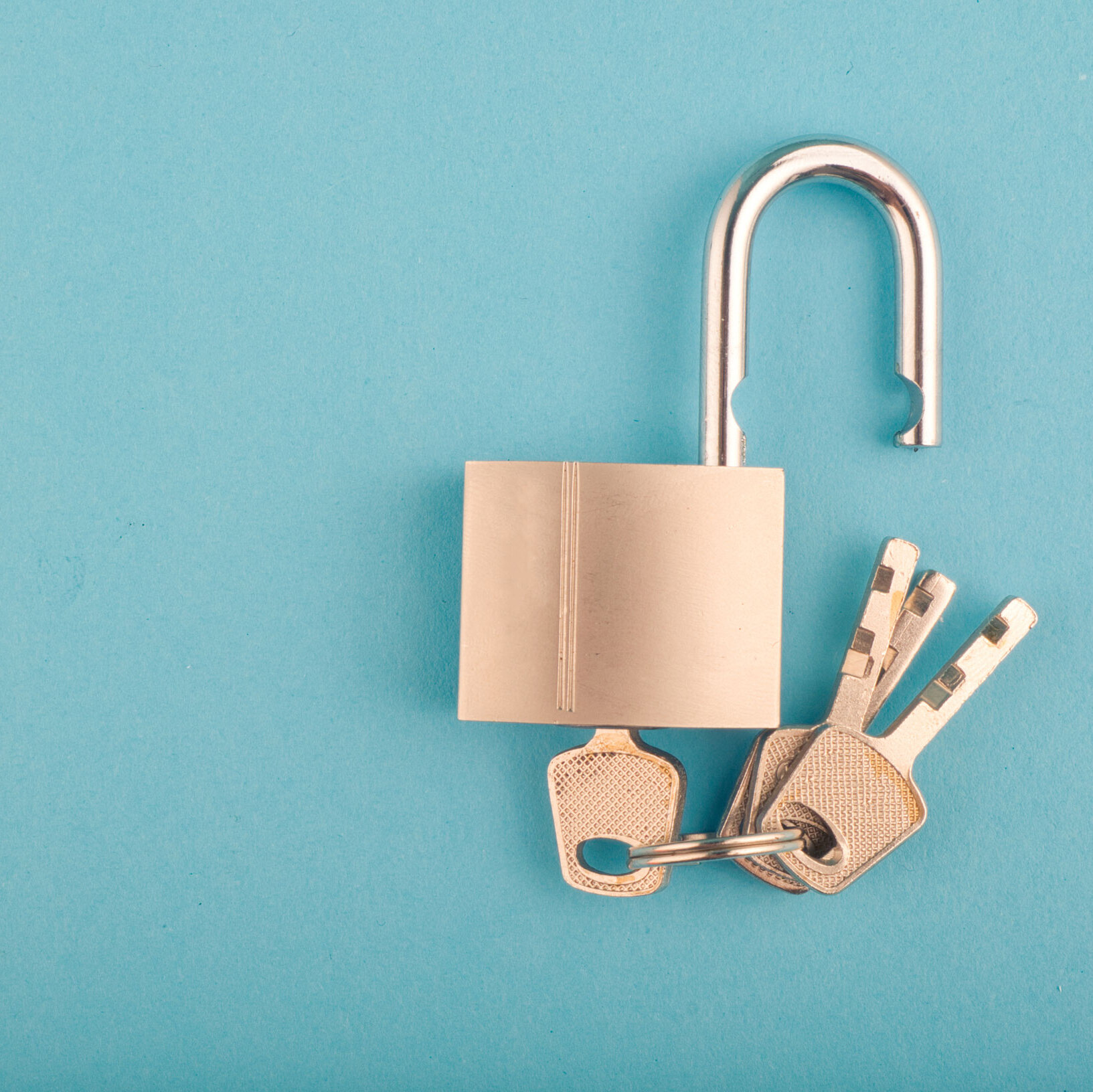Ein silbernes Vorhängeschloss mit geöffnetem Bügel liegt auf einer hellblauen Fläche; am Schlossring hängen drei silberne Schlüssel, die nebeneinander angeordnet sind.