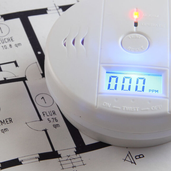 Ein Kohlenmonoxidmelder liegt auf einem Wohnungsgrundriss. Das Display zeigt einen Messwert von 0 und eine kleine Kontrollleuchte ist aktiviert.