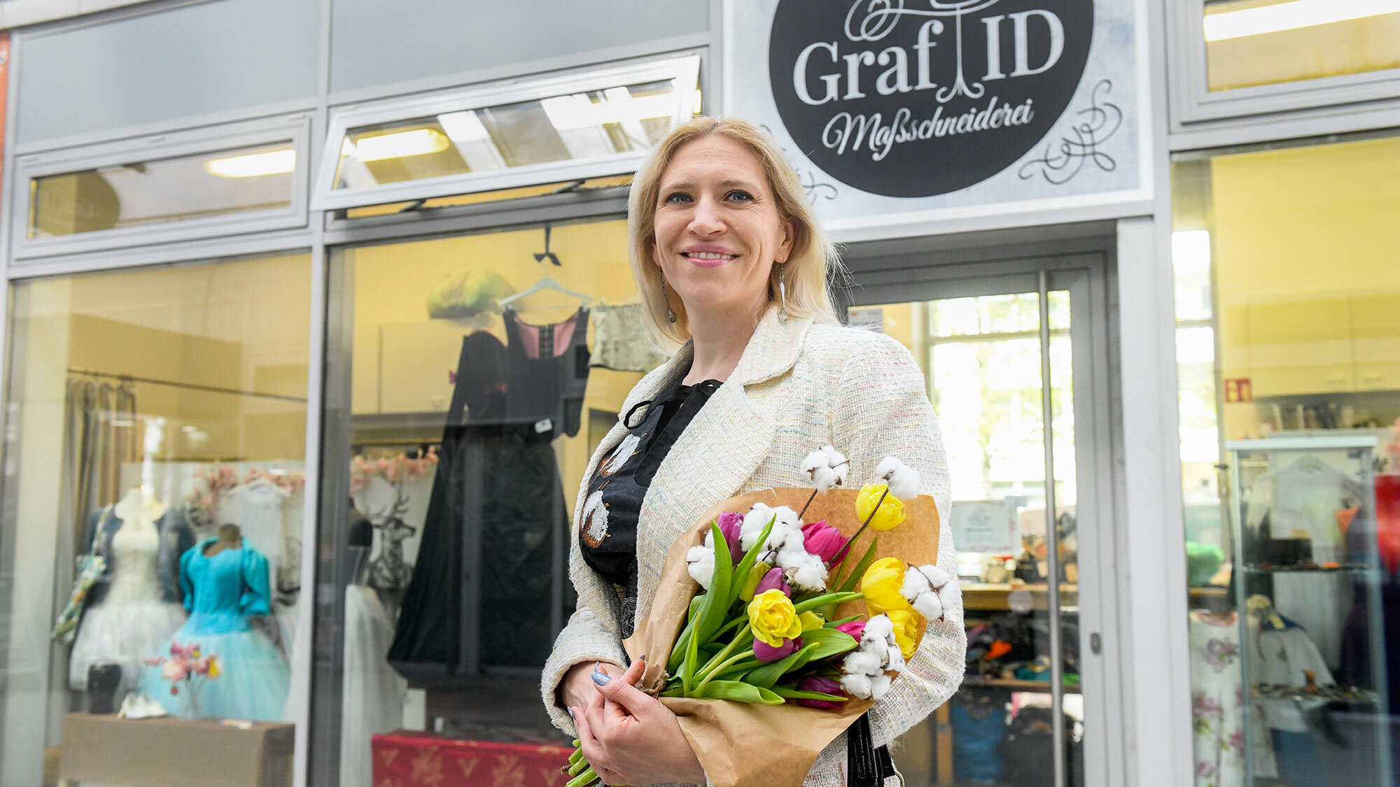 Die Schneiderin Darya Graf steht vor ihrem Atelier auf der Marzahner Promenade. Sie hält einen Blumenstrauss in den Armen und lächelt in die Kamera. Im Schaufenster des Ateliers sind Kostüme zu erkennen.
