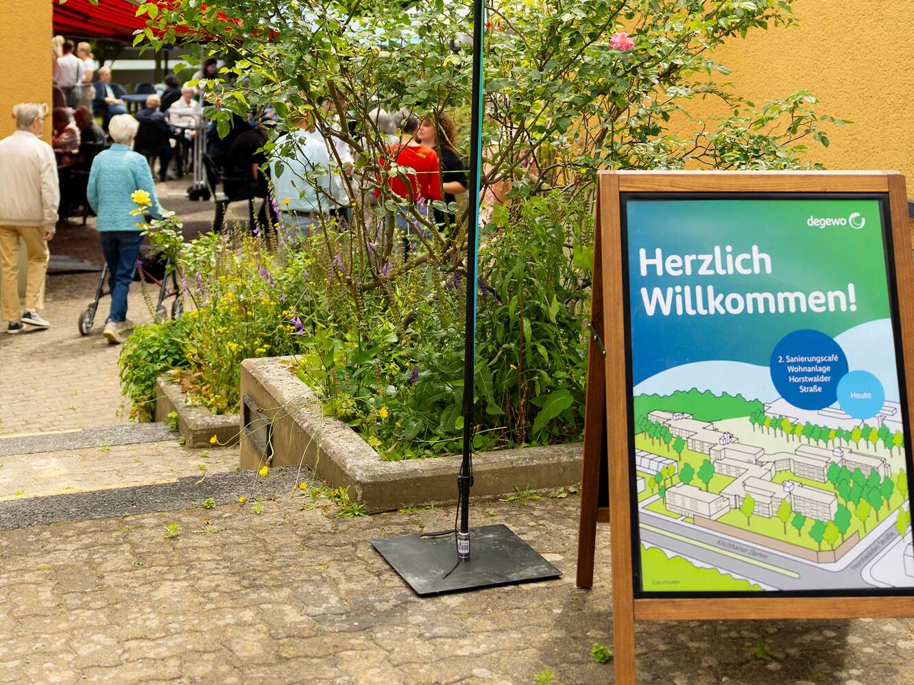 Willkommenstafel zum Sanierungscafé der Wohnanlage Horstwalder Straße steht im Vordergrund, im Hintergrund treffen sich Bewohnende im Außenbereich.