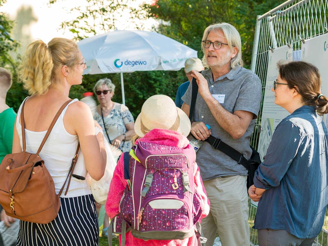 Kleingruppe von Anwohnenden im Gespräch vor Projektinformationen am Bauzaun. Ein Mitarbeiter erläutert Inhalte, während Erwachsene und Kinder zuhören; ein degewo-Sonnenschirm steht im Hintergrund.