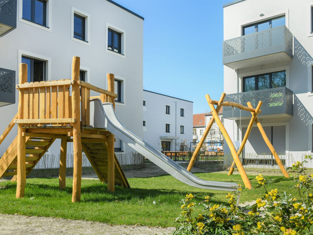 Außenansicht degewo-Neubau Rudower Straße 47 bis 51 mit Spielplatz und blühendem Busch im Vordergrund.