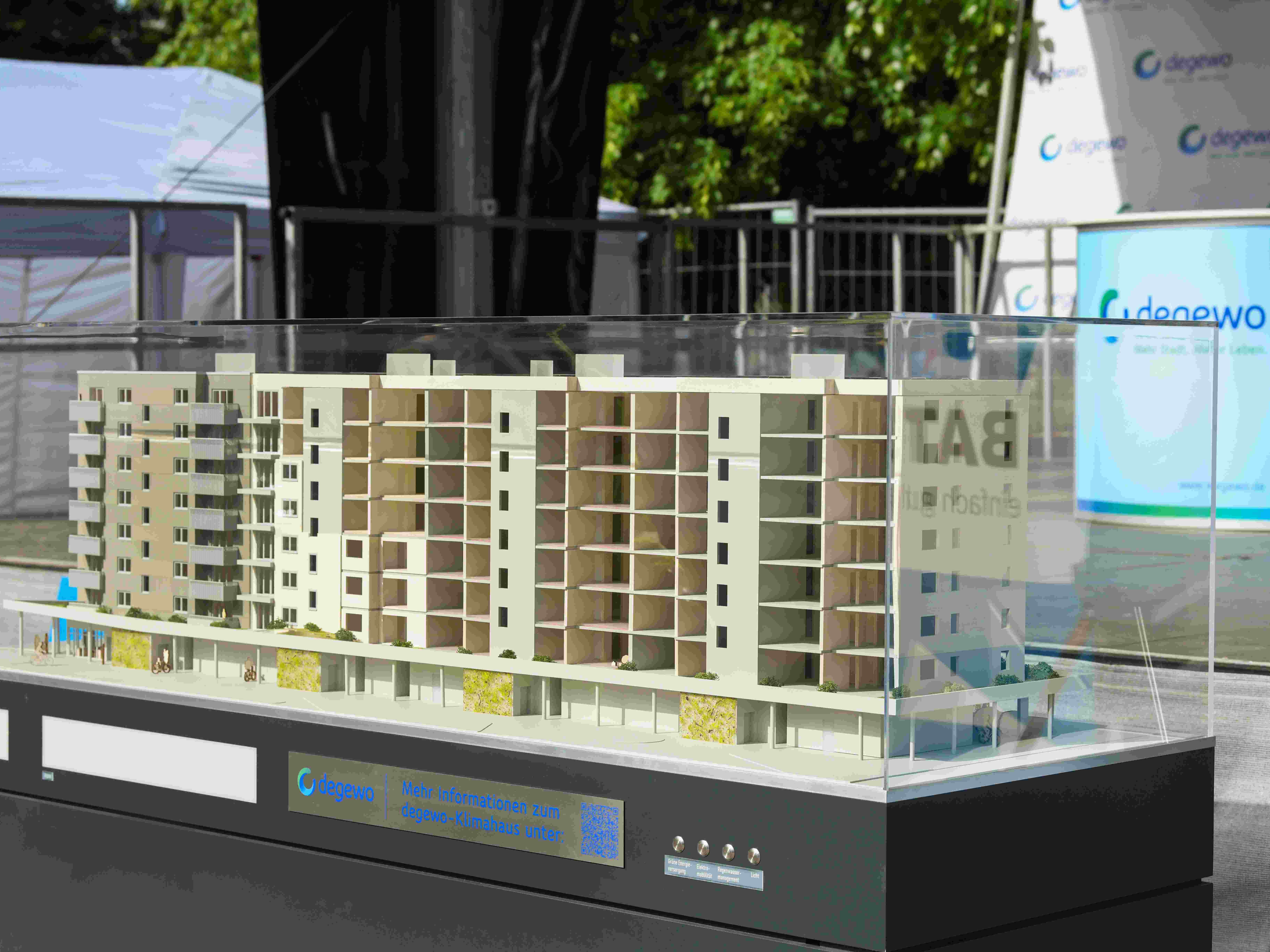 Ein detailliertes Modell eines mehrgeschossigen Wohngebäudes steht in einer transparenten Vitrine auf einem dunklen Sockel mit degewo-Beschriftung. Das Modell zeigt Balkone, Fassadenstruktur und eine Sockelzone mit Gewerbeflächen. Im Hintergrund befinden sich Pavillons und eine degewo-Infowand.