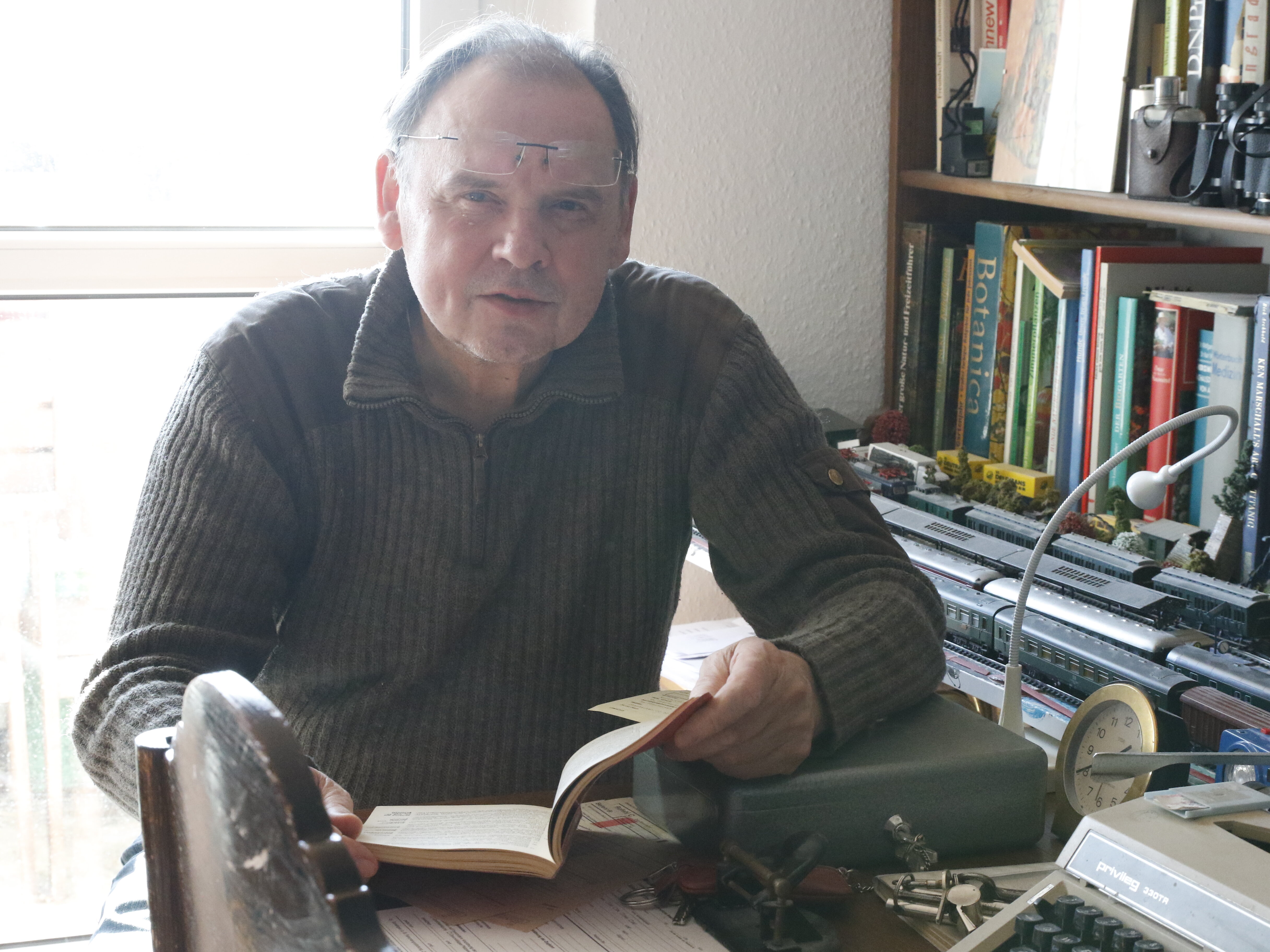 Mann sitzt in einem Zimmer an einem Schreibtisch, hinter ihm ist ein Fenster und Bücherregal zu sehen. Er hat ein Buch aufgeschlagen und blickt in die Kamera.