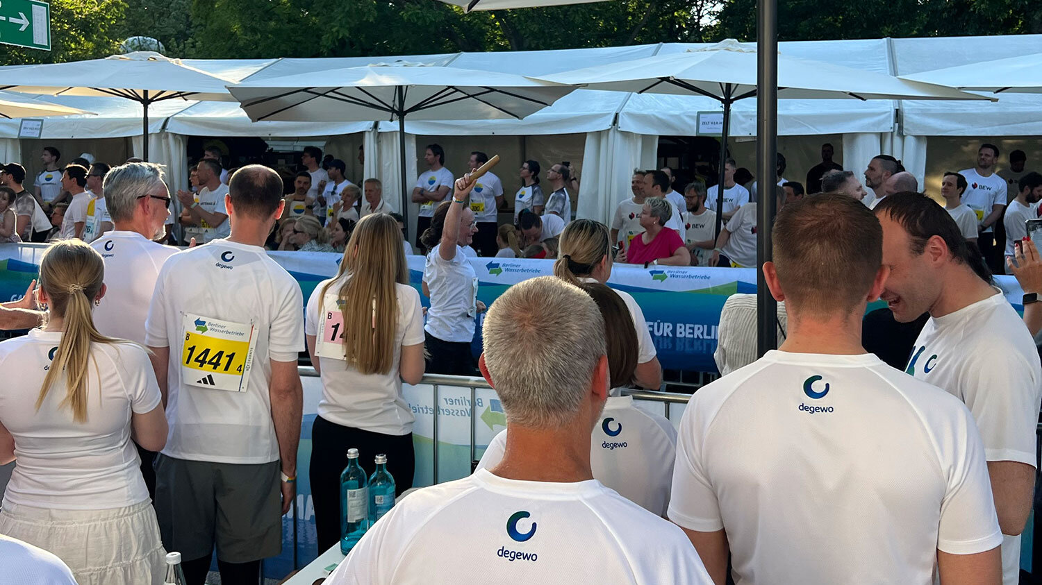 Das Foto zeigt Läufer bei einem Teamstaffellauf in Berlin. Sie stehen mit dem Rücken zur Kamera und haben ein Logo von degewo auf dem Laufshirt. Sie stehen hinter einer Bande und jubeln den einlaufenden Kolleginnen und Kollegen zu.