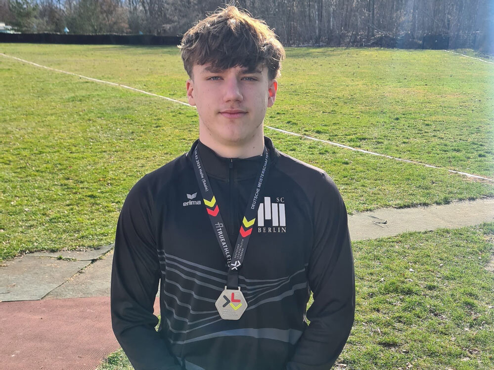 Junger Athlet steht auf einem Rasenplatz und trägt eine Medaille um den Hals. Er blickt ruhig in die Kamera.