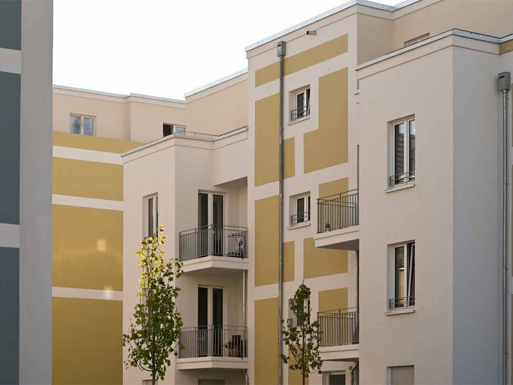 Nahaufnahme der Fassade eines degewo-Neubaus am Eisenhutweg mit Balkonen und gelben Akzentflächen. Junge Bäume und Grünflächen ergänzen die moderne Architektur und unterstreichen den wohnlichen Charakter der neu errichteten Wohnanlage.