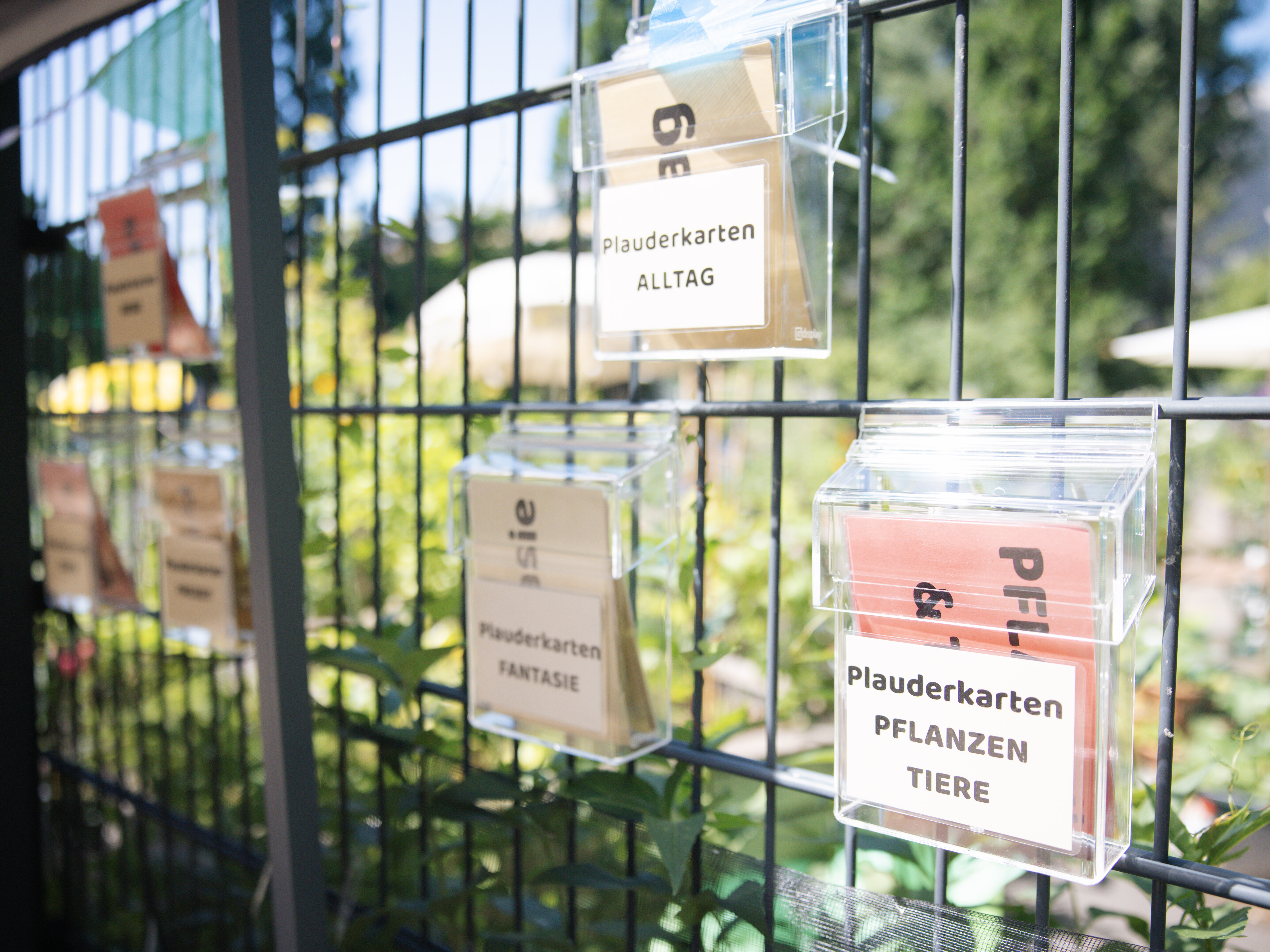 Kästen mit Plauderkarten hängen an einem Zaun. Auf den Karten stehen Begriffe wie "Alltag", "Pflanzen und Tiere" und "Fantasie". 