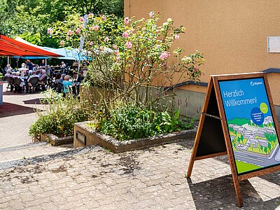 Ein Aufsteller von degewo mit der Aufschrift Willkommen steht vor einem Gebäude. Im Hintergrund ist eine Informationsveranstaltung zu sehen, die durch viele Anwohnenden besucht ist. Sie sitzen unter Zelten.