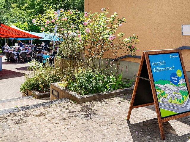 Ein Aufsteller von degewo mit der Aufschrift Willkommen steht vor einem Gebäude. Im Hintergrund ist eine Informationsveranstaltung zu sehen, die durch viele Anwohnenden besucht ist. Sie sitzen unter Zelten.