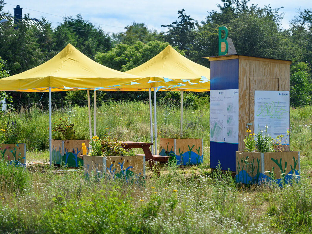 Gelbe Pavillons mit Sitzgelegenheiten und Infotafeln auf einer Wiese. Der Stand ist Teil einer Partizipationsaktion, umgeben von bepflanzten Holzkästen.