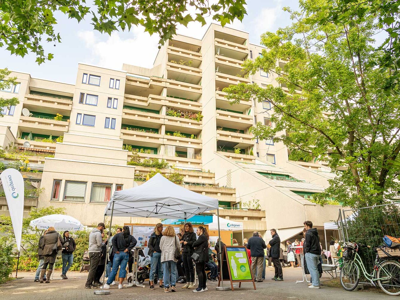 Außenaufnahme der Schöneberger Terrassen in Berlin-Schöneberg während einer degewo-Informationsveranstaltung zur Sanierung. Vor dem terrassenförmig angelegten Wohngebäude stehen Pavillons und Infotafeln, mehrere Mieterinnen und Mieter kommen ins Gespräch. Begrünte Balkone und Bäume rahmen die Szene.