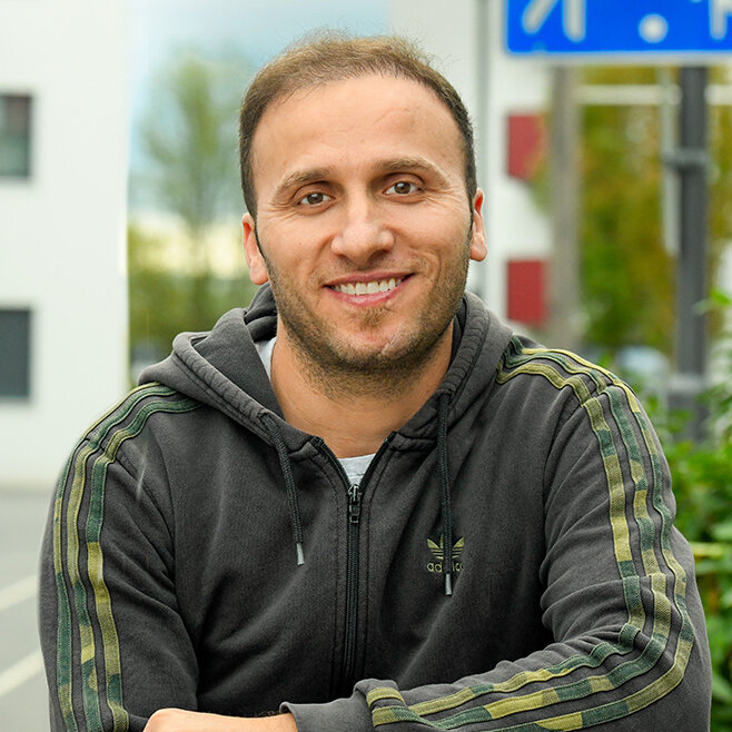 Mann steht mit verschränkten Armen im Außenbereich einer Wohnanlage, im Hintergrund sind spielende Kinder, Wohngebäude und Grünflächen zu sehen; links im Bild das Logo „Wir sind degewo“.