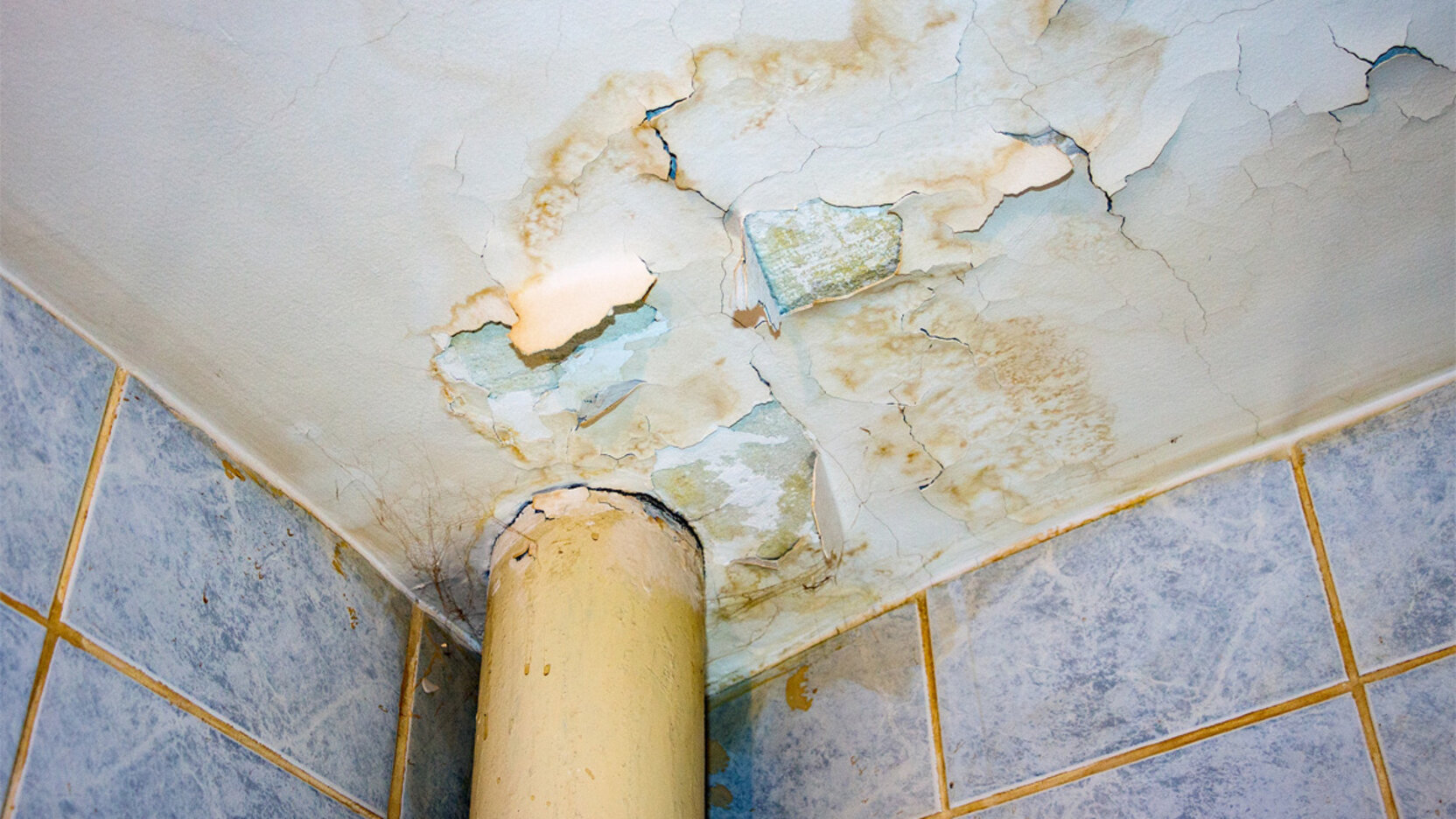 Decke eines Badezimmers mit sichtbaren Feuchtigkeitsflecken, abblätternder Farbe und Rissen rund um ein Rohr; darunter sind geflieste Wände zu sehen.