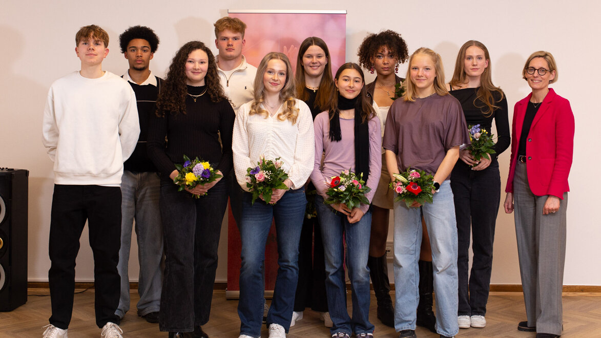 Gruppenfoto mehrerer junger Athletinnen und Athleten, die in einem Innenraum nebeneinander stehen und Blumensträuße halten. Rechts steht eine erwachsene Person in einem roten Blazer.