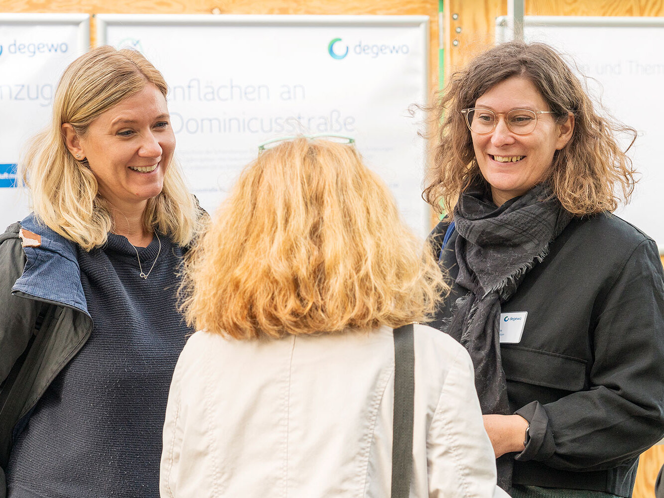 Zwei degewo-Mitarbeiterinnen im persönlichen Gespräch mit einer Besucherin vor einer Infotafel zur Sanierung und zu den Ladenflächen an der Dominicusstraße. 