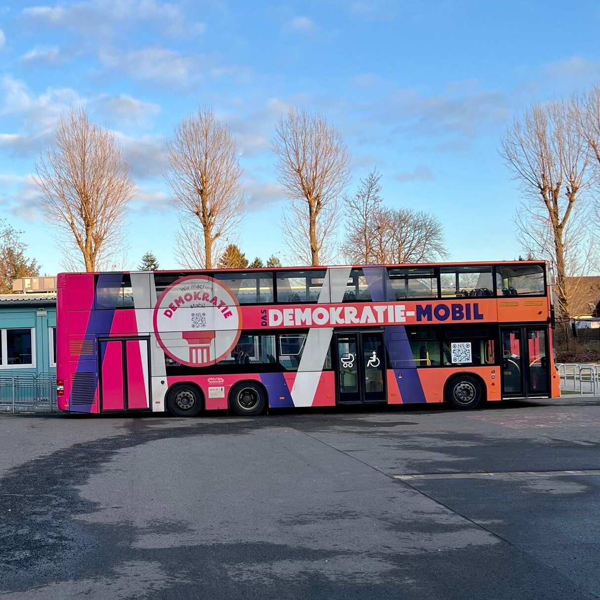 Bunt gestalteter Doppeldeckerbus "Demokratie-Mobil" steht vor Gebäuden und Bäumen.
