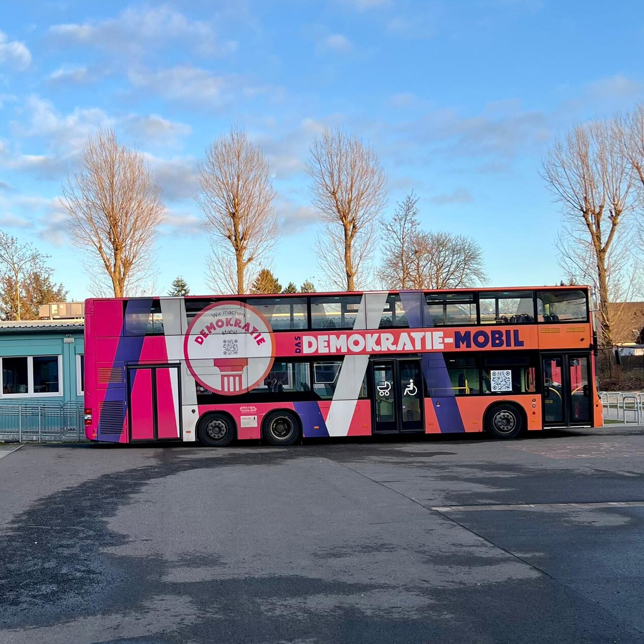 Bunt gestalteter Doppeldeckerbus "Demokratie-Mobil" steht vor Gebäuden und Bäumen.