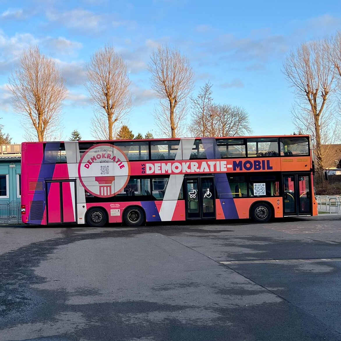 Bunt gestalteter Doppeldeckerbus "Demokratie-Mobil" steht vor Gebäuden und Bäumen.