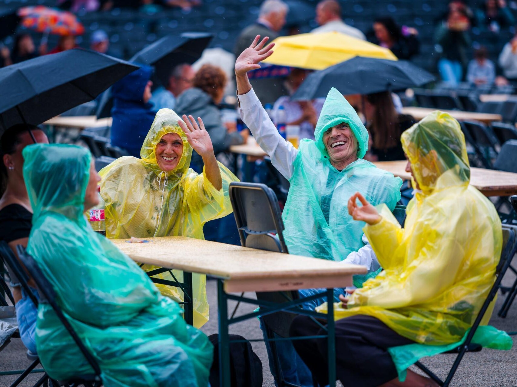 Mehrere Besucherinnen und Besucher sitzen bei leichtem Regen an Tischen im Freien. Sie tragen farbige Regenponchos und winken in die Kamera, während im Hintergrund weitere Gäste unter Regenschirmen sitzen.