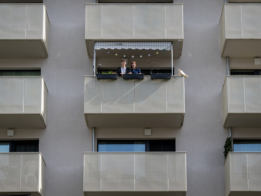 Zwei Menschen stehen auf einem mittleren Balkon eines modernen Wohnhauses und blicken lächelnd nach unten.