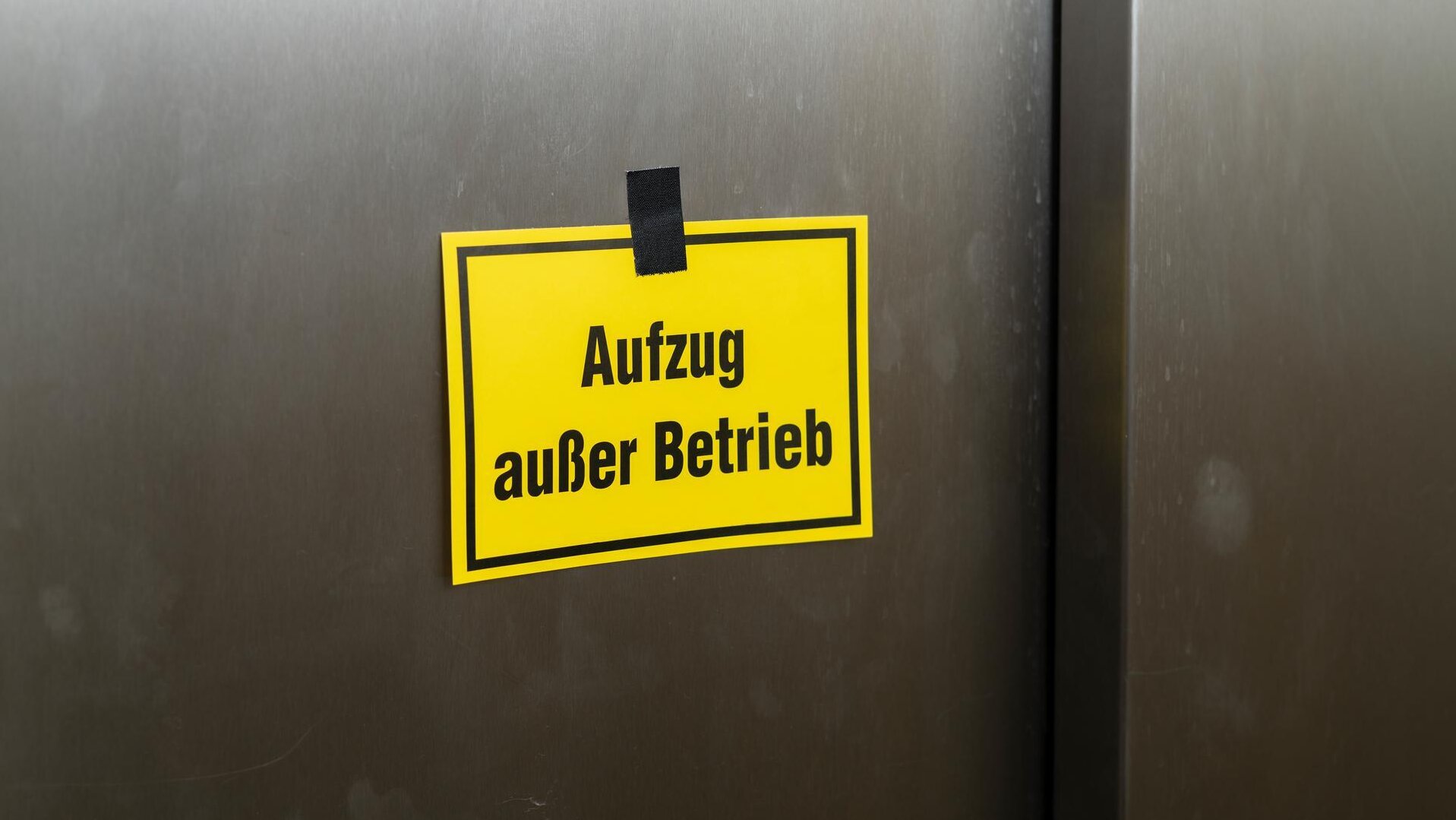 Gelbes Schild mit der Aufschrift „Aufzug außer Betrieb“, mit schwarzem Klebeband an einer dunklen Aufzugstür befestigt.