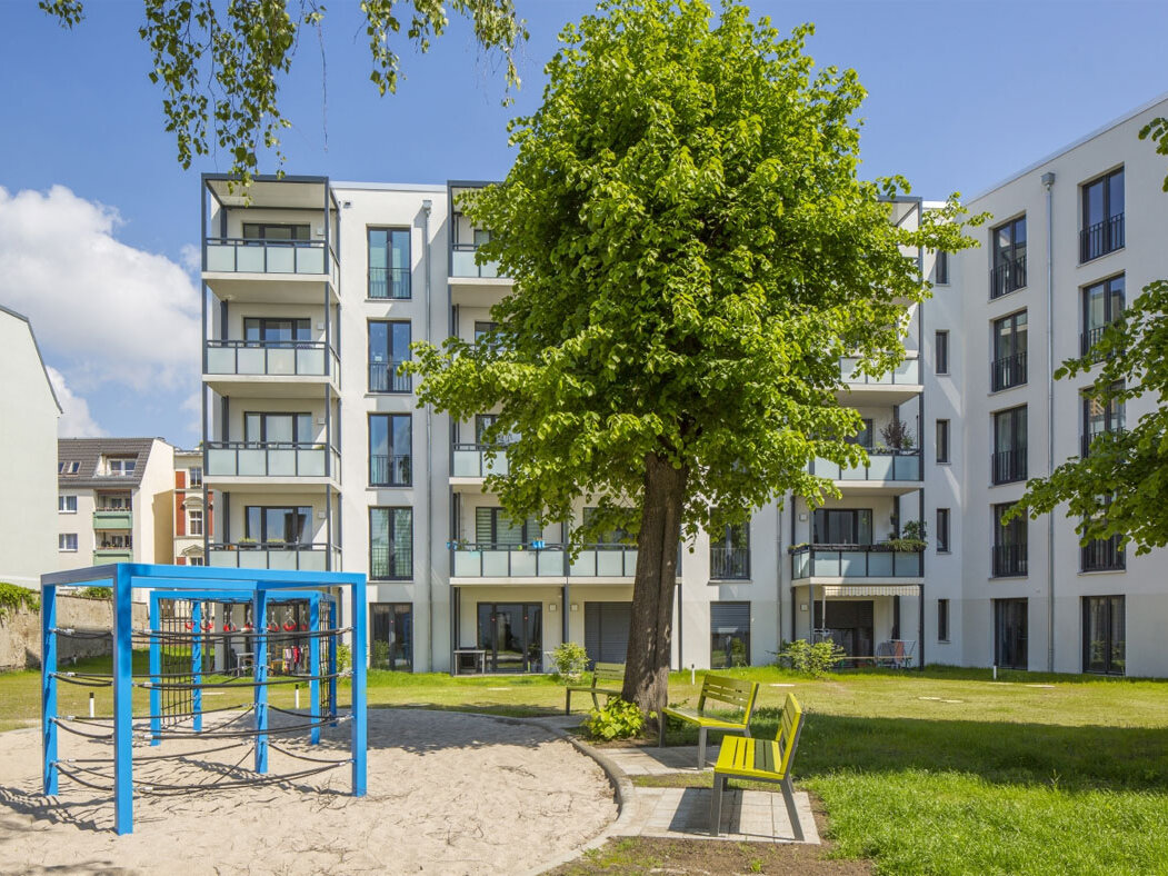 Außenansicht degewo-Neubau Luisenstraße 24 bis 24 A: Hofseite mit Spielplatz, Sitzbänken, Grünfläche und Bäumen im Vordergrund.