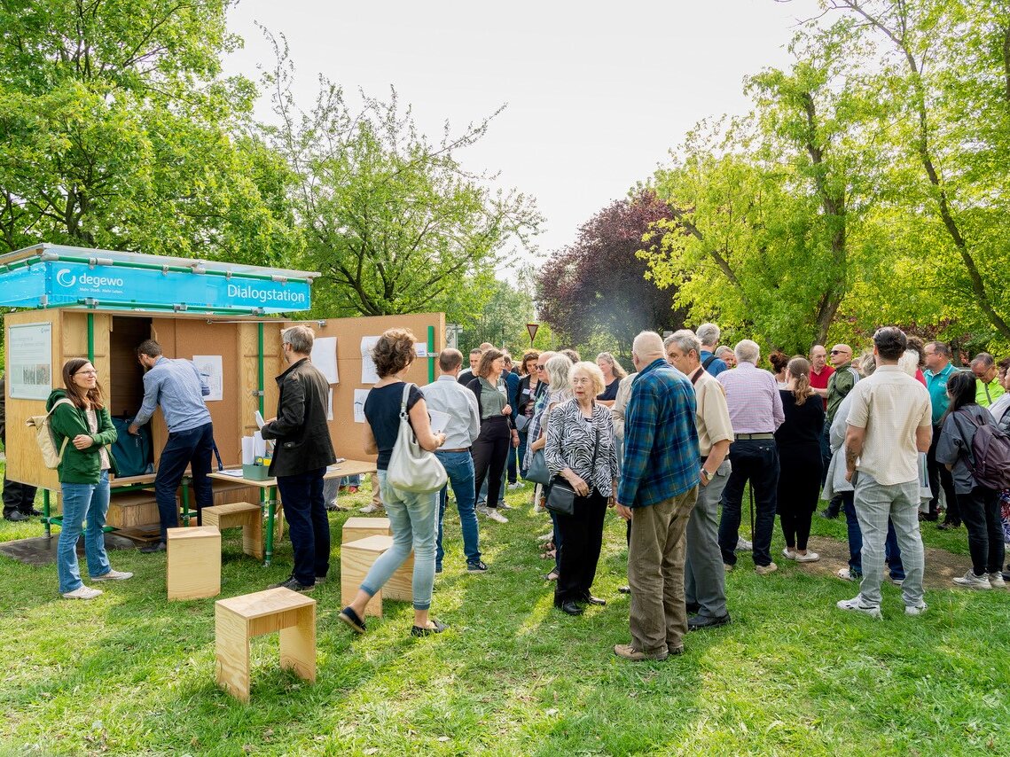 Gesprächsrunde mit Anwohnenden im Freien; ein Moderator erklärt Inhalte, während Teilnehmende aufmerksam zuhören.
