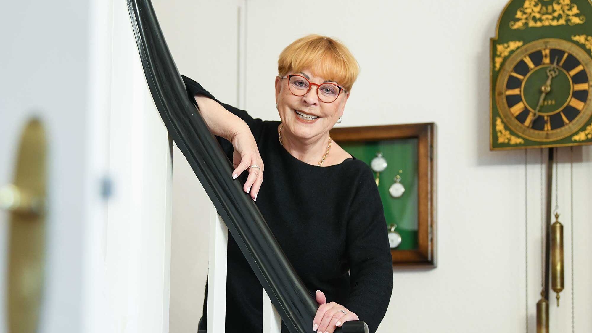 Eine ältere Frau lehnt sich entspannt an ein Treppengeländer in ihrer Wohnung. Hinter ihr hängen alte Uhren und gerahmte Sammlerstücke an der Wand.