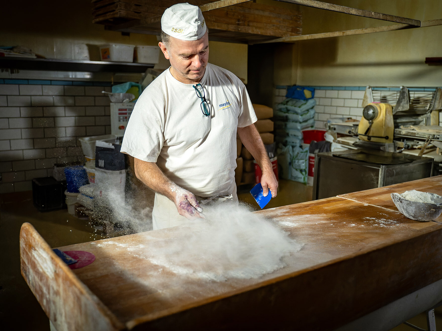 Bäcker in weißer Arbeitskleidung streut Mehl über einen Holztisch in einer Backstube mit Geräten und Mehlsäcken im Hintergrund.