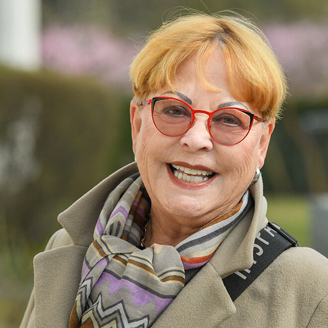 Eine ältere Frau mit roter Brille lächelt in die Kamera. Sie trägt einen Mantel und einen bunten Schal.