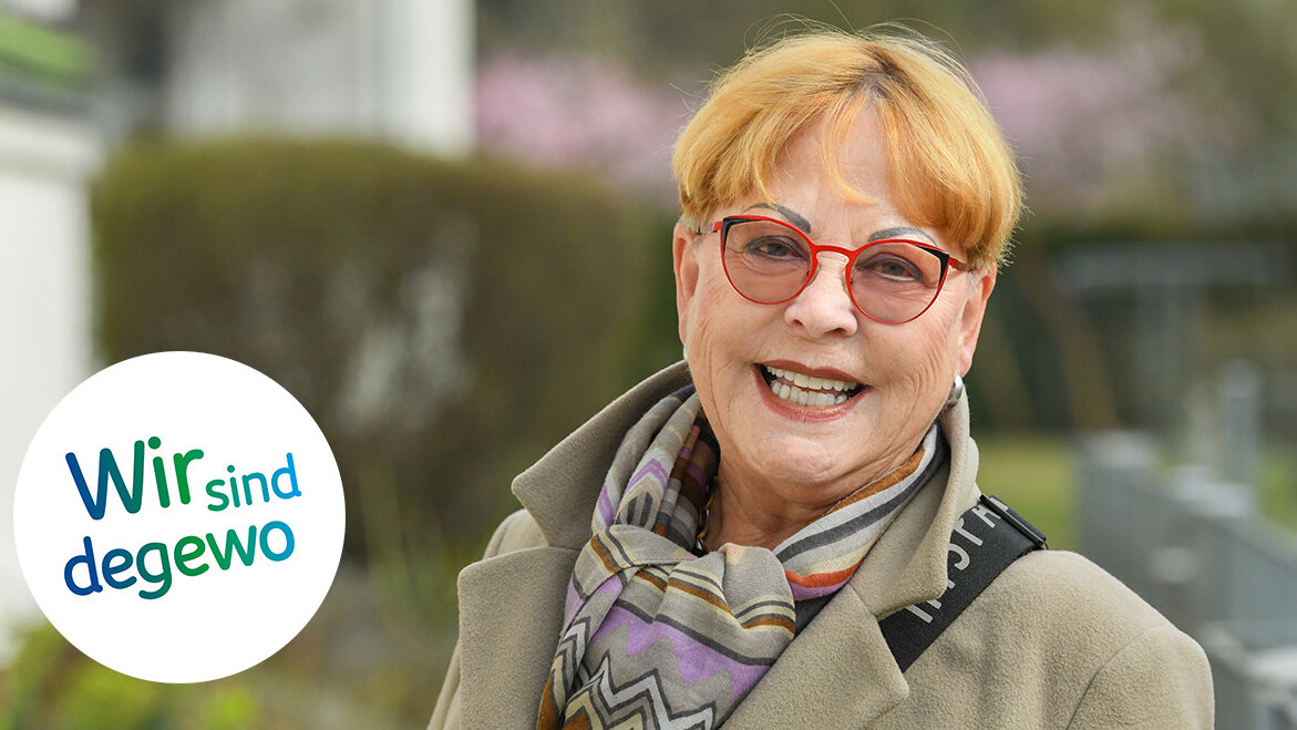 Eine ältere Frau mit roter Brille lächelt in die Kamera. Sie trägt einen Mantel und einen bunten Schal.