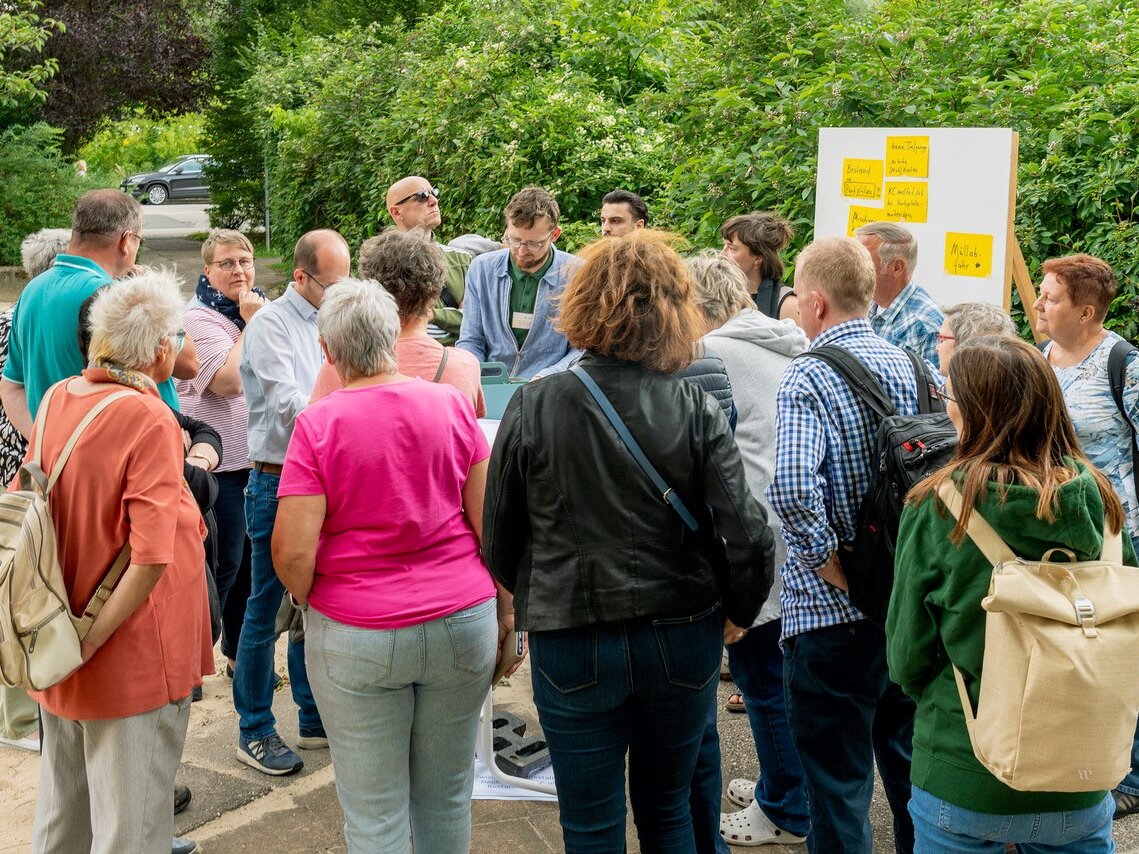 Große Gruppe von Anwohnenden diskutiert um einen Tisch; im Hintergrund eine Pinnwand mit Haftnotizen.