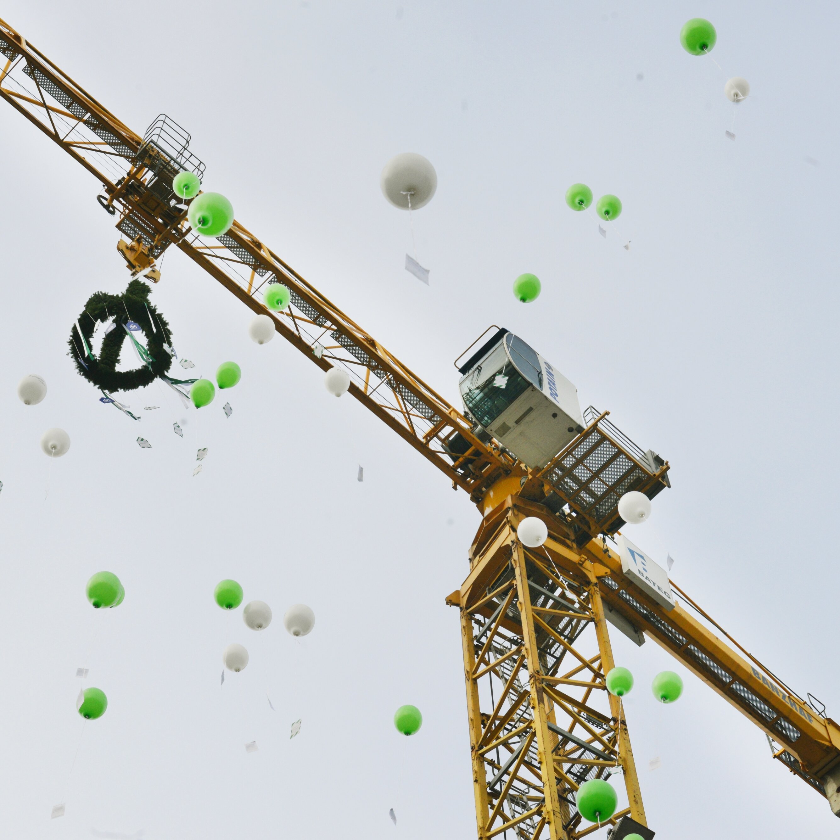 Aufnahme eines Baukrans vor grauem Himmel. Um den Kran steigen grüne und weiße Luftballons auf, teilweise mit angehängten Karten. Das Motiv vermittelt eine feierliche Stimmung, beispielsweise im Zusammenhang mit einem Richtfest oder Projektabschluss.