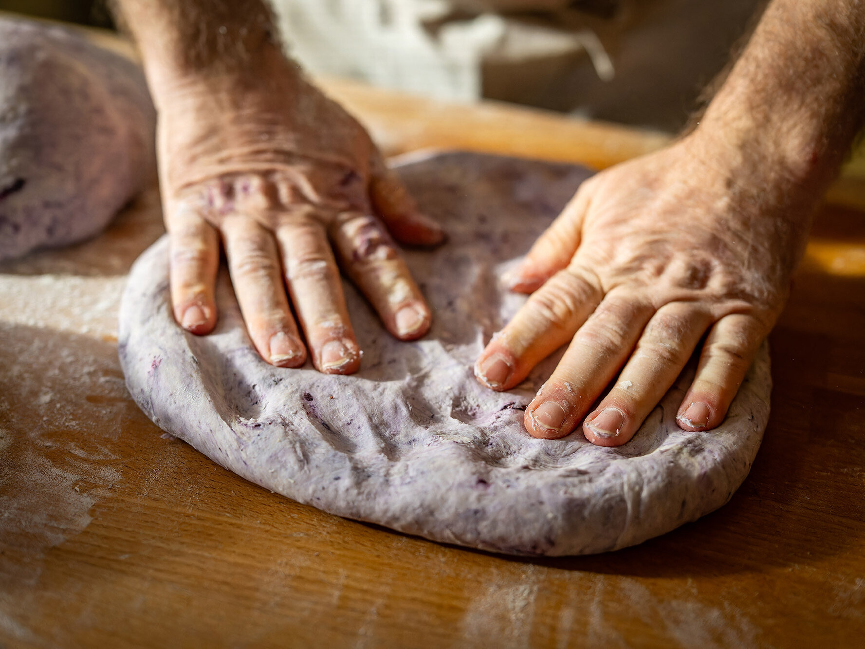In einer Nahaufnahme sind mehlbedeckte Hände kneten violetten Teig auf einem hölzernen Arbeitstisch zu sehen.