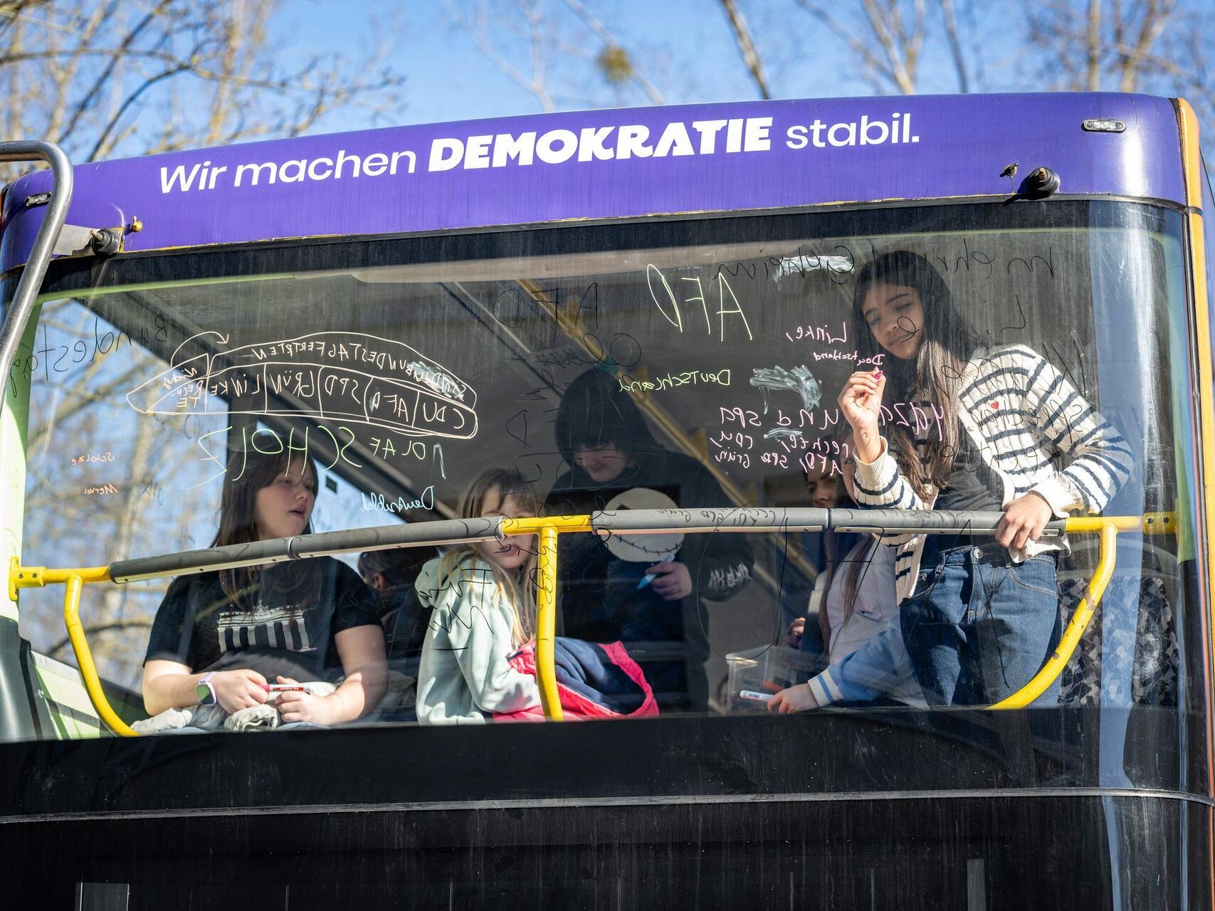 Mehrere Kinder stehen und sitzen hinter der Frontscheibe des Demokratie-Mobils und schreiben etwas mit Fensterstiften an die Scheibe.