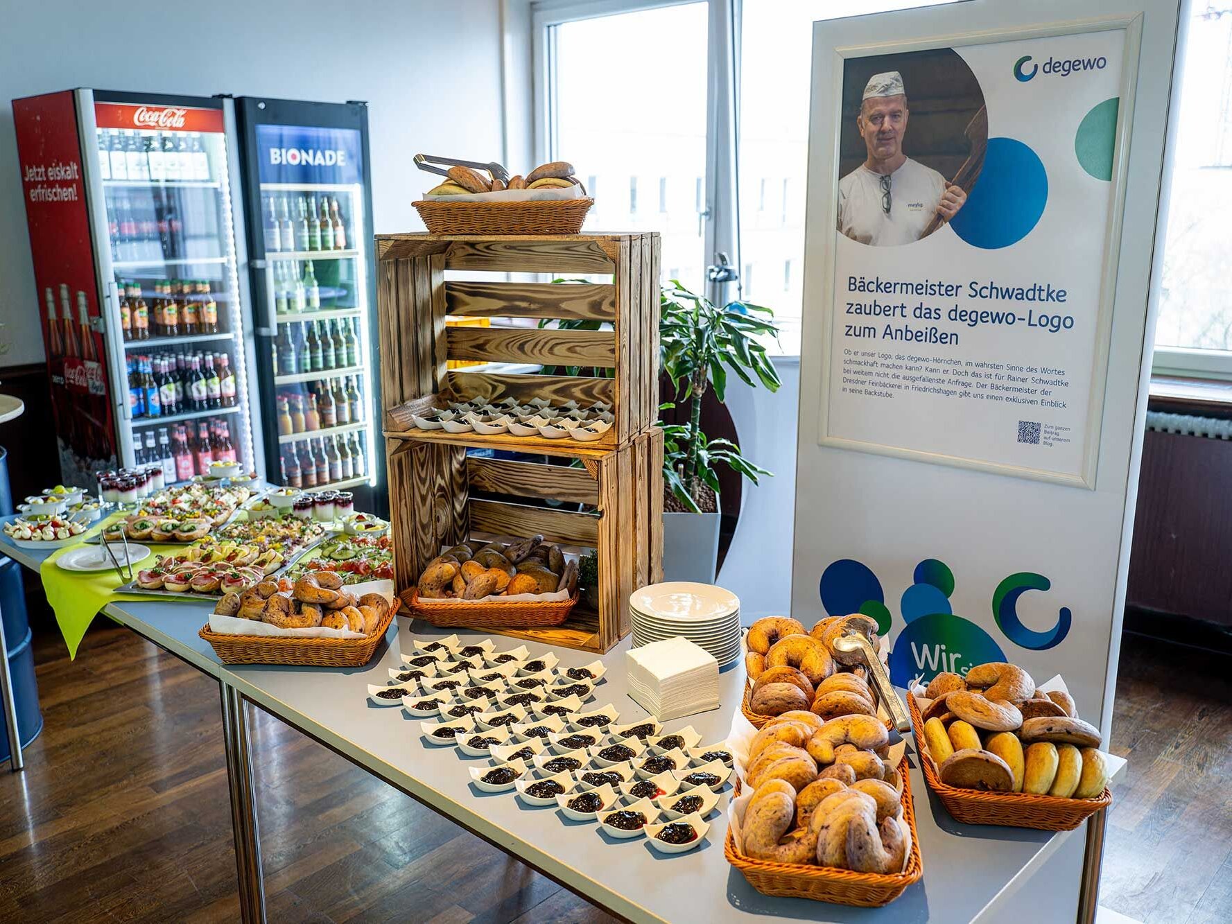 Ein Buffet mit Gebäck belegten Brötchen und kleinen Desserts ist aufgebaut während ein Plakat über einen Bäckermeister und das degewo Logo informiert.
