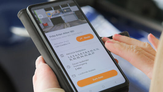 Zwei Hände halten ein Smartphone, auf dessen Bildschirm eine Seite der Parkhaus-App PAYUCA zu sehen ist.