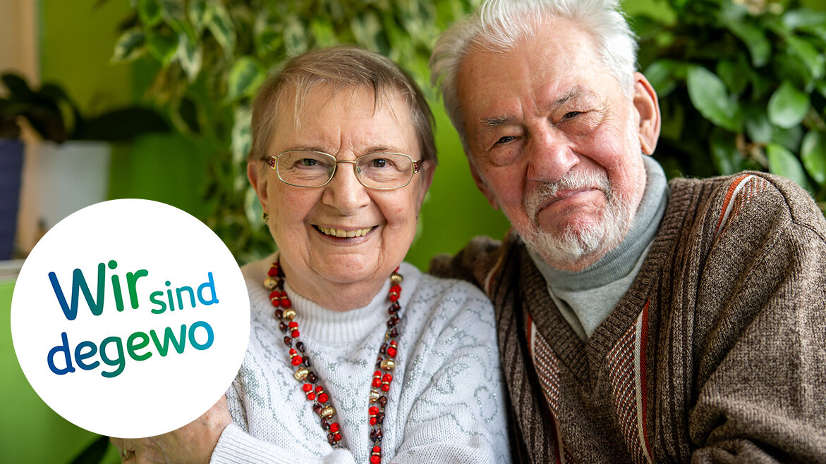 Ein älteres Paar sitzt eng umschlungen vor grüner Zimmerpflanzenkulisse und lächelt warm in die Kamera.