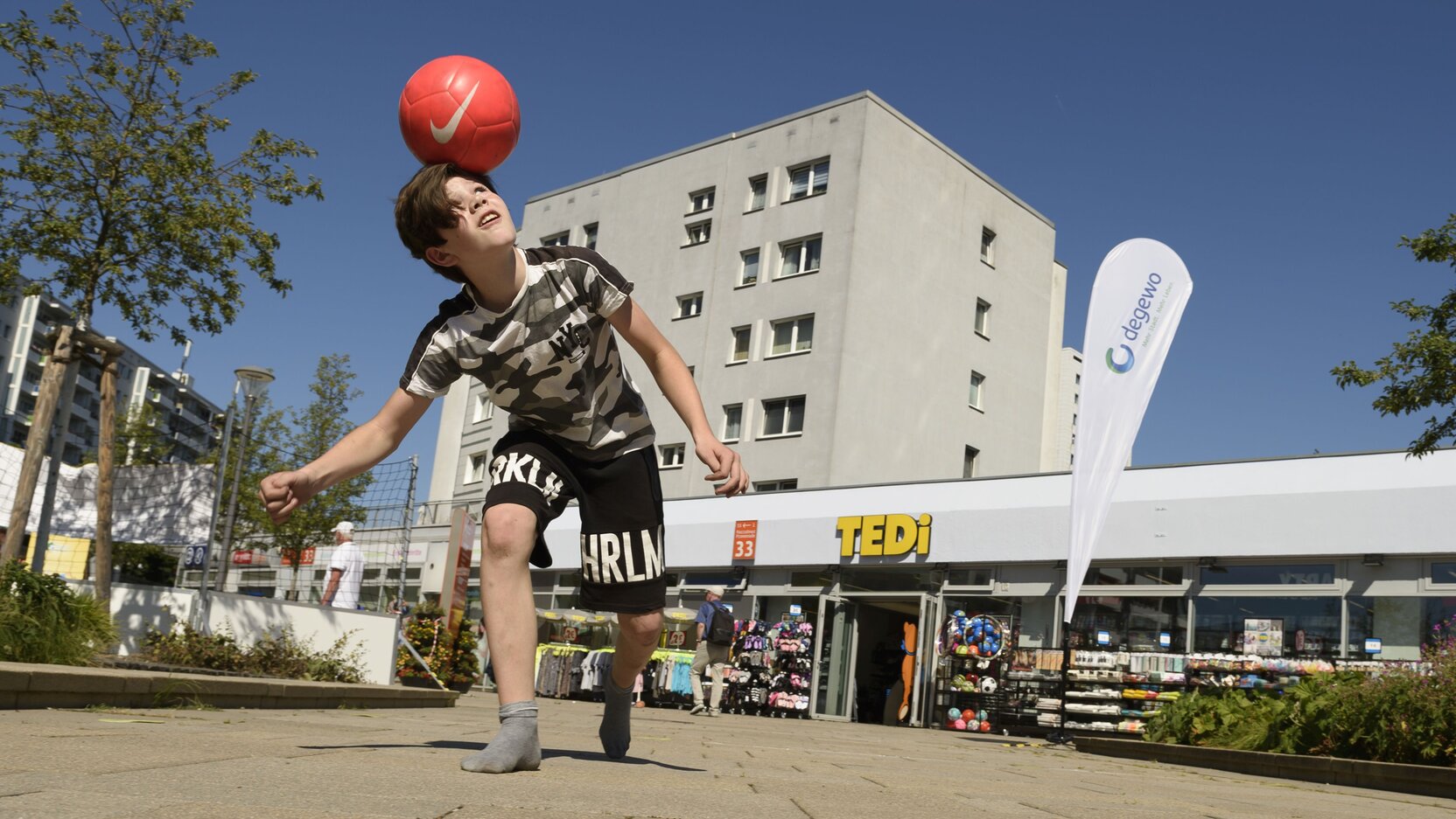 Ein Junge balanciert vor einer Ladenzeile einen Fußballball auf dem Kopf.