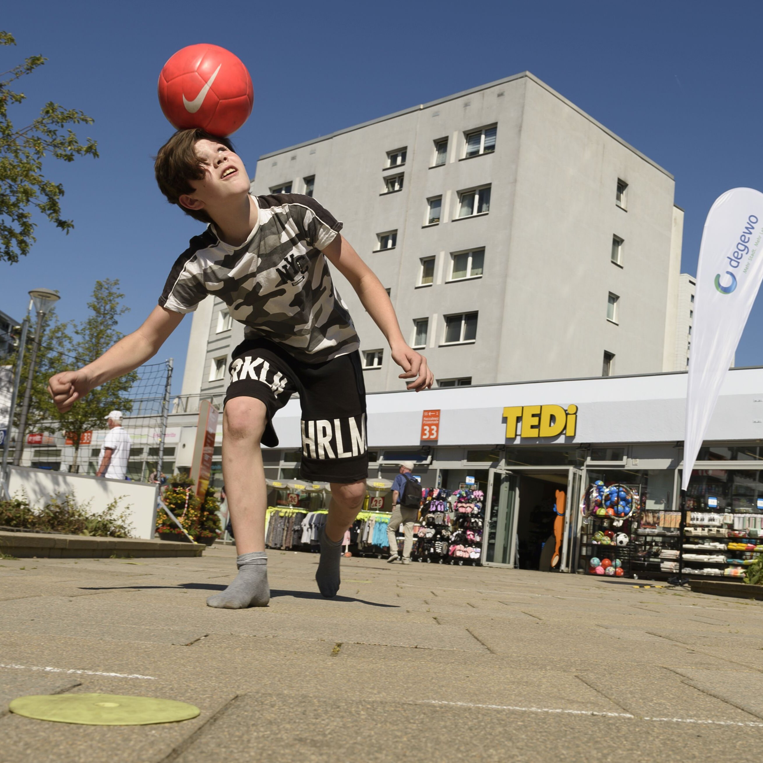 Ein Junge balanciert vor einer Ladenzeile einen Fußballball auf dem Kopf.