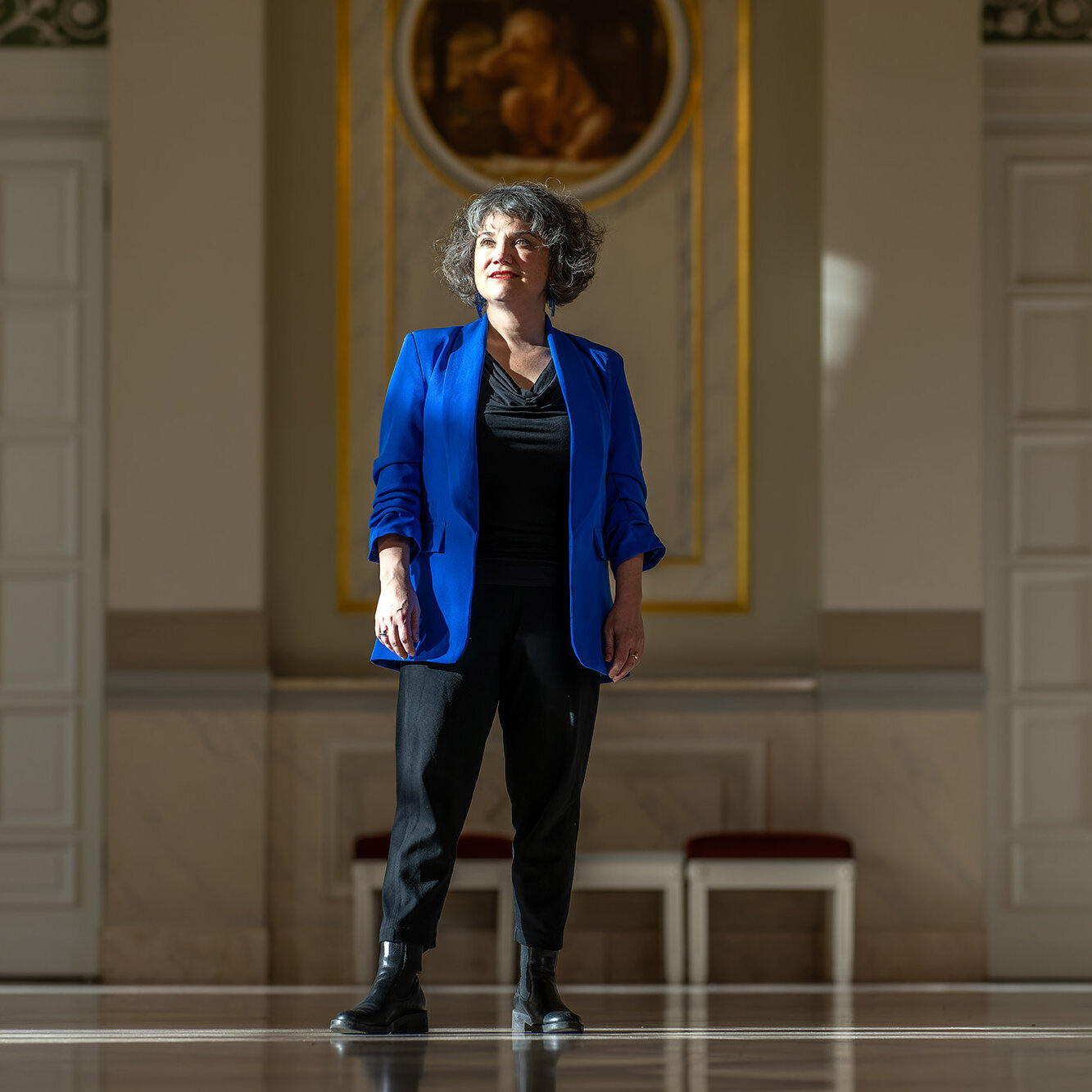 Eine Frau steht in einem großen historischen Saal und trägt ein blaues Sakko. Sie blickt leicht nach oben und wird vom Sonnenlicht angestrahlt.