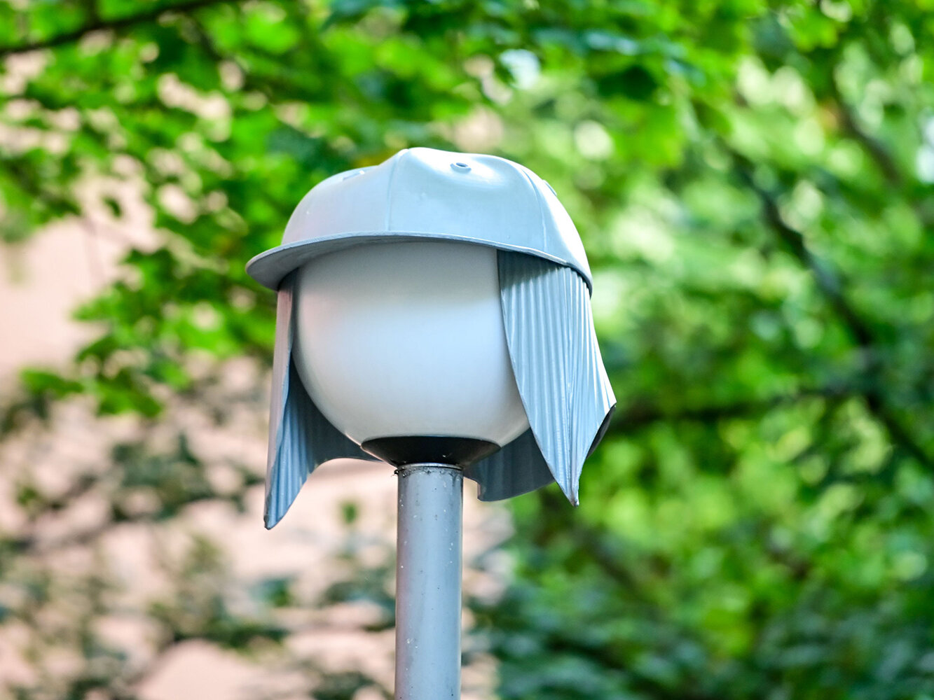 Eine runde Straßenleuchte ist mit einer grauen Kappe verkleidet. Das Lichtobjekt steht vor grünen, dichten Baumkronen.