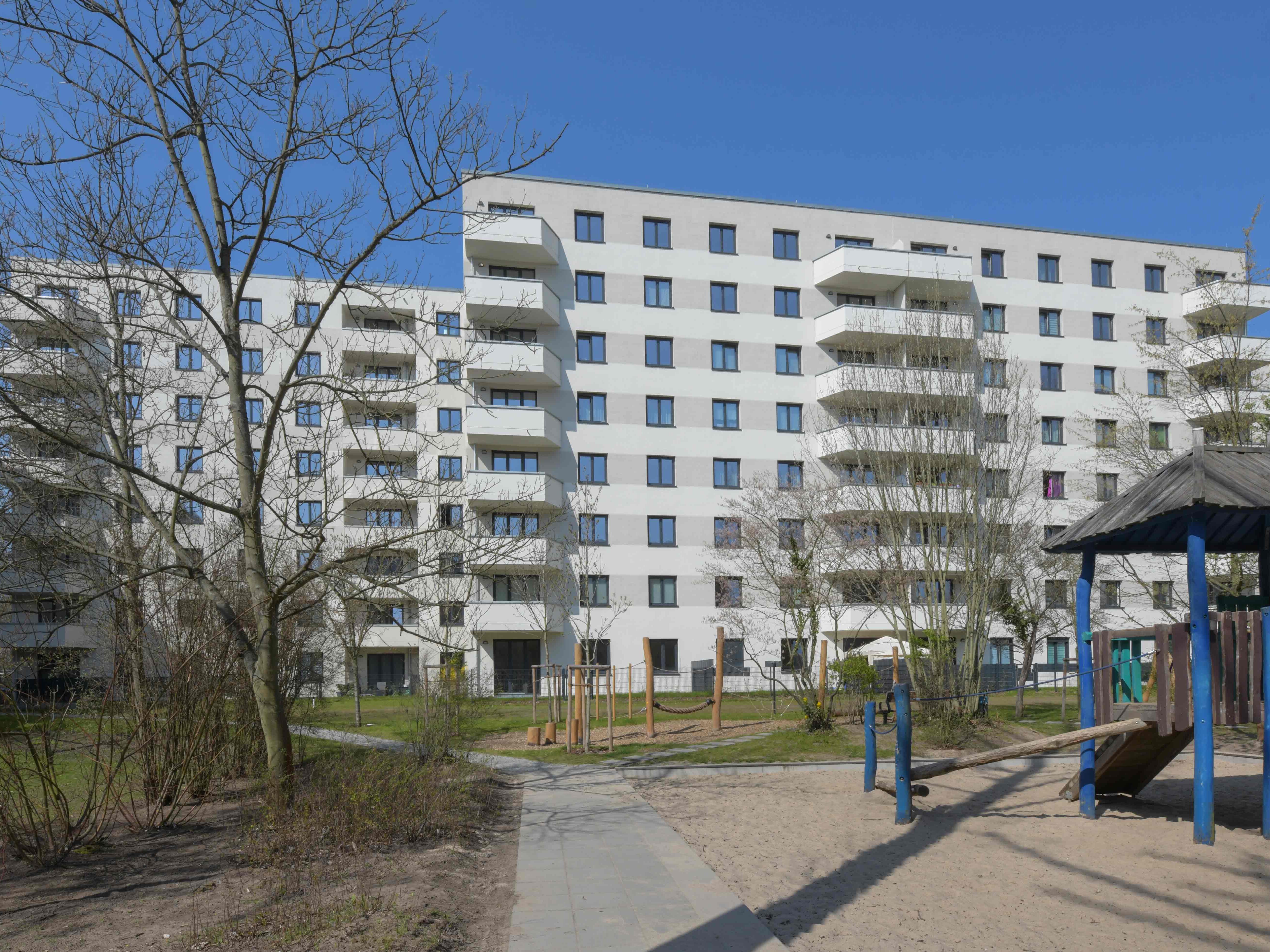 Außenansicht degewo-Neubau Wuhlestraße 2 bis 8 mit Bäumen, Weg und Spielplatz im Vordergrund.