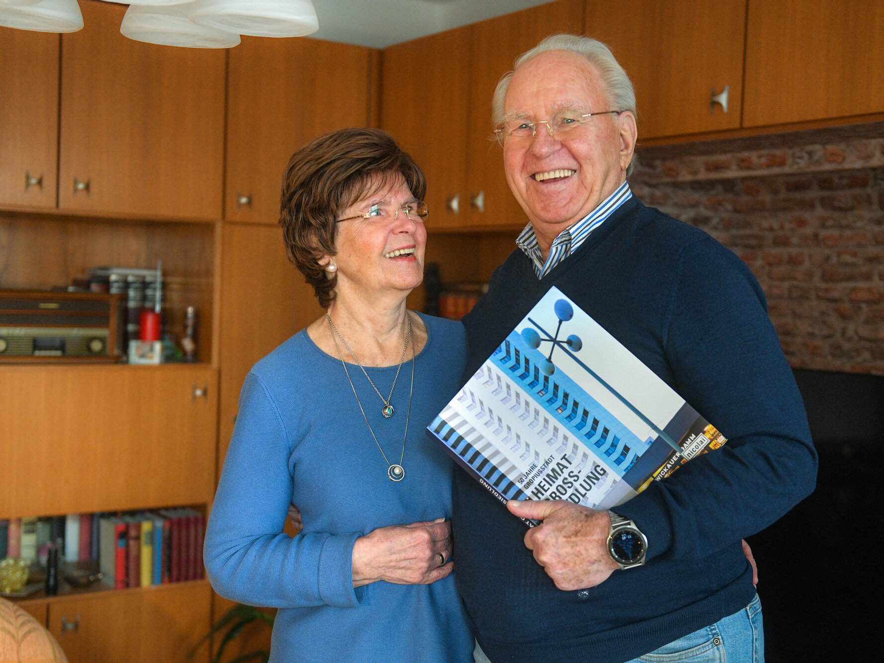 Ein älteres Paar lächelt in einer gemütlichen Wohnung; der Mann hält ein Buch mit historischem Foto eines Hochhauses.