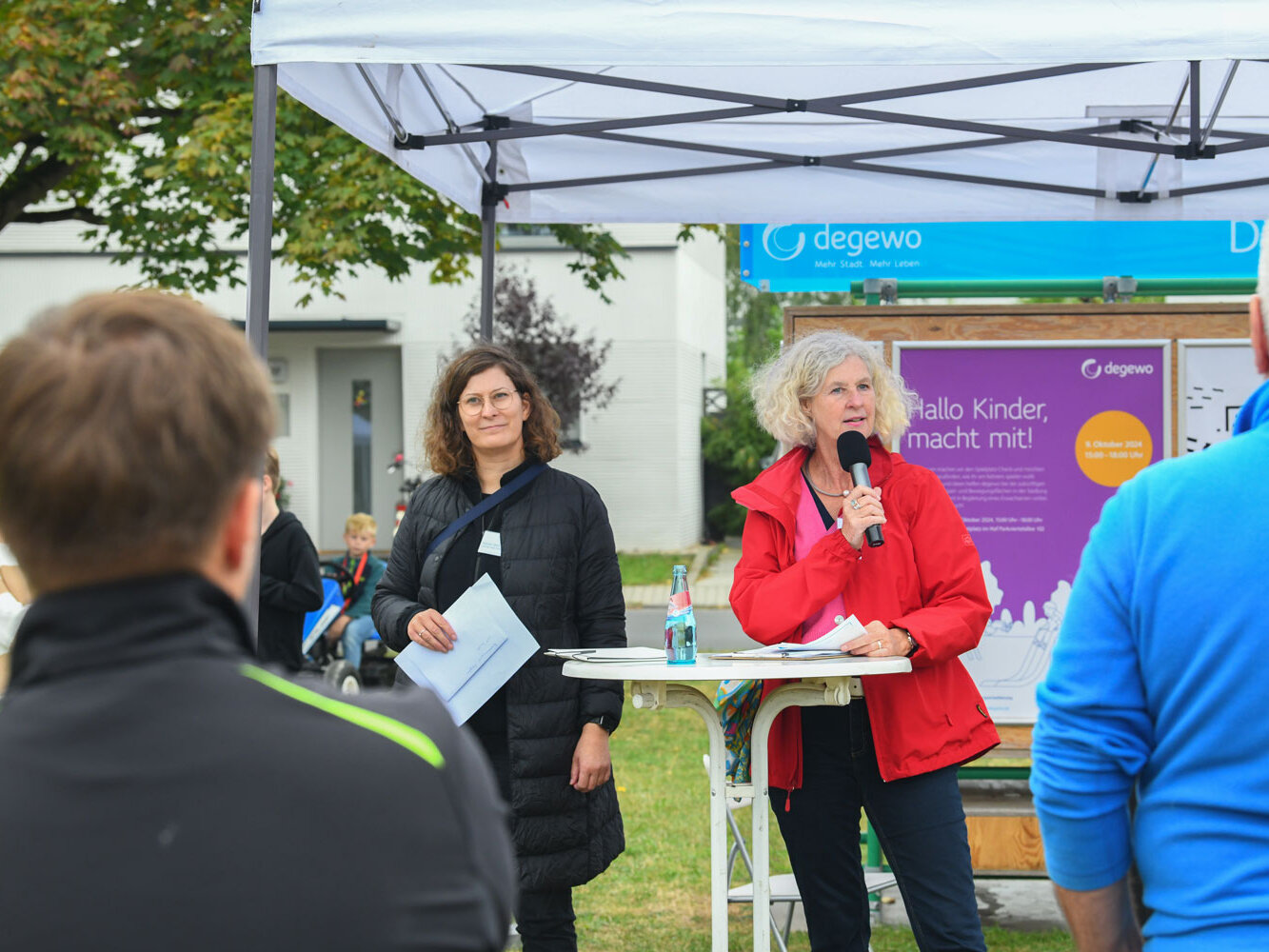 Eine Frau mit Mikrofon in der Hand steht an einem Stehtisch, neben ihr eine weiter Personen. Im Hintergrund eine Wand mit Plakaten.