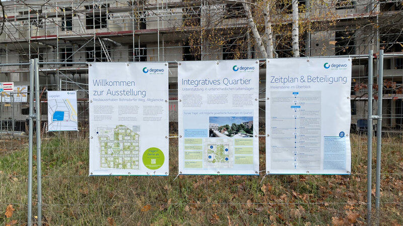 Mehrere Informationstafeln zum Neubauvorhaben Bohnsdorfer Weg hängen an einem Bauzaun. Im Hintergrund ist ein Gebäude im Bau mit Gerüst zu sehen, davor herbstliche Vegetation.
